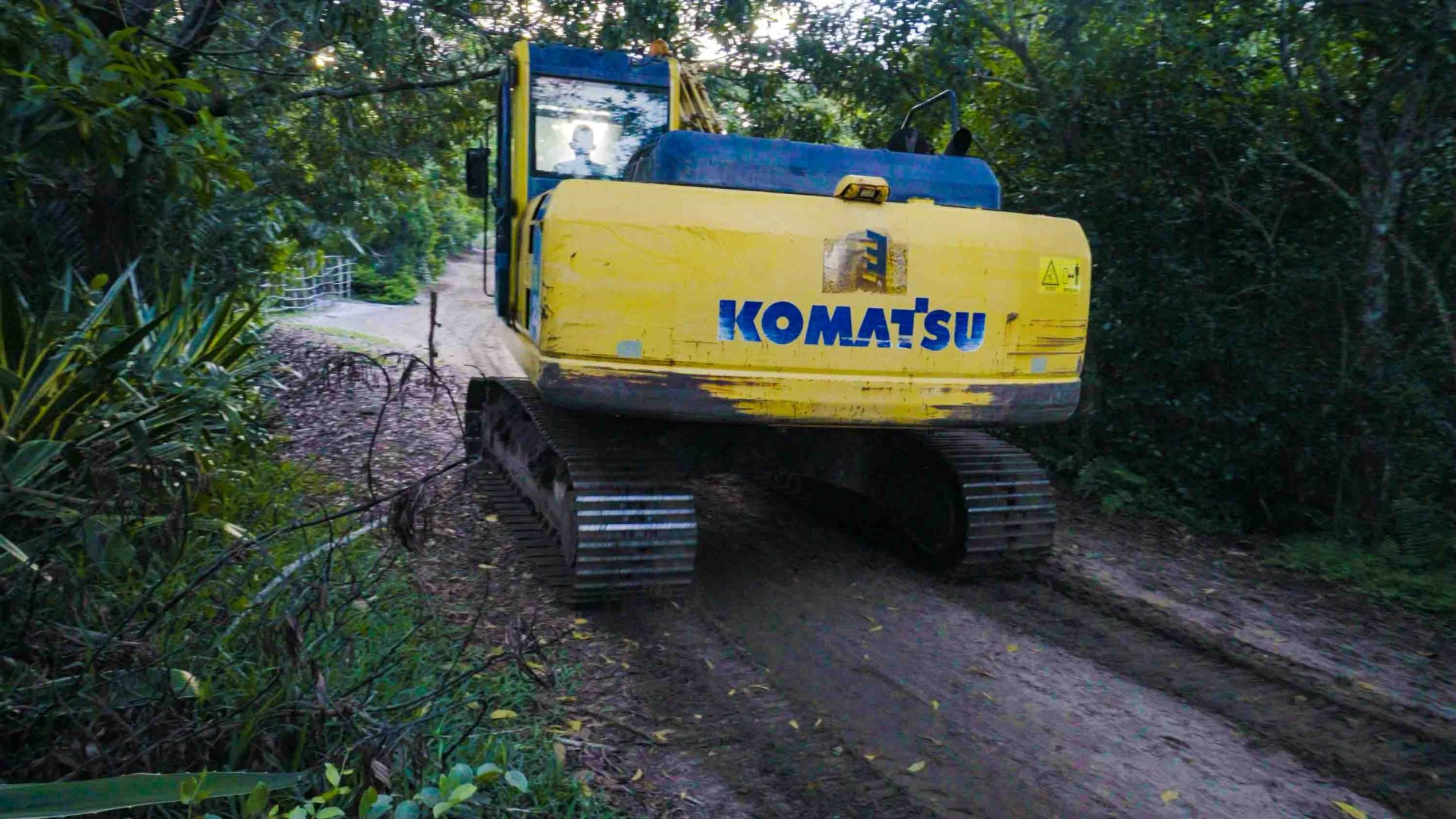 A yellow and blue piece of construction equipment drives on a dirt road through forest.