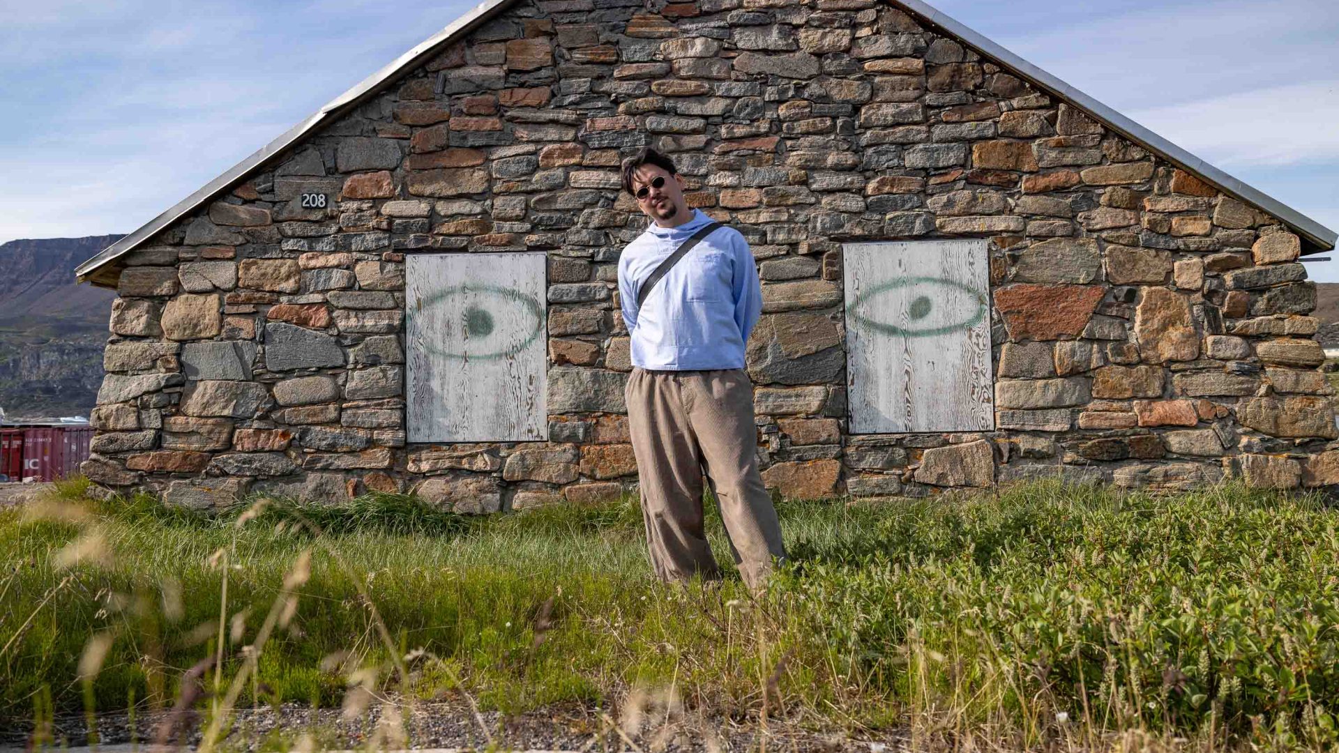 A man in brown trousers and a white top outside a stone house.