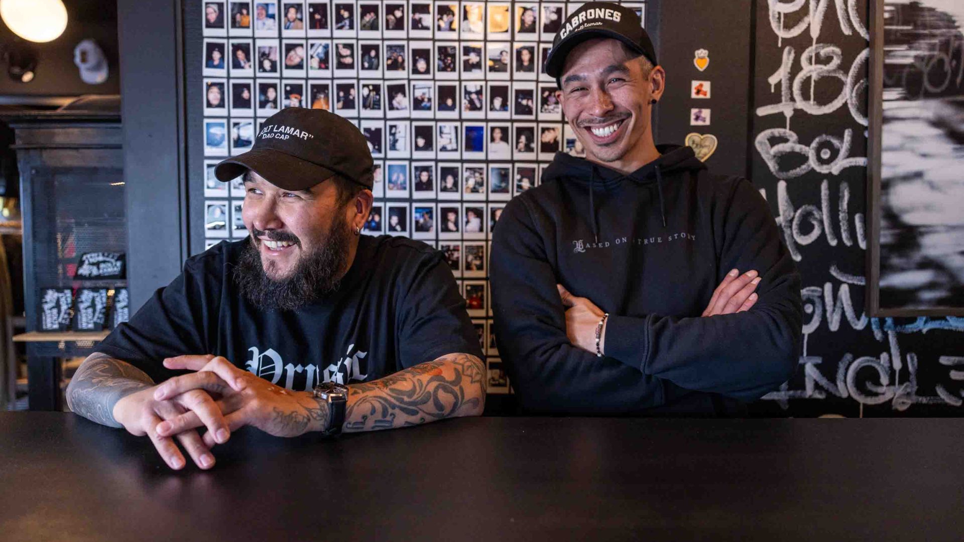 Two men in black streetwear and caps smile to camera at a shop counter