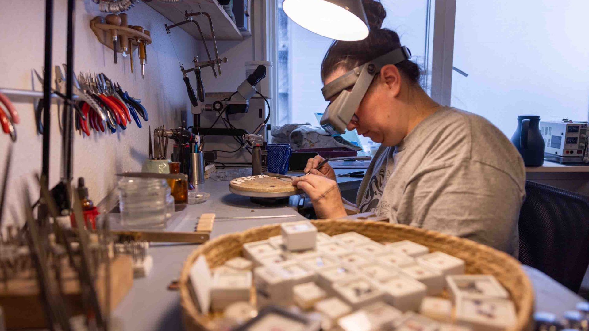 A woman in protective eye wear works on making jewellery in a workshop. She has a trayof pieces next to her.