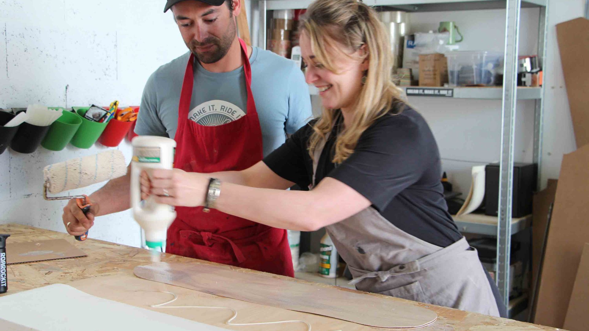 A woman applied glue to the board of a skateboard that she is making with a man.