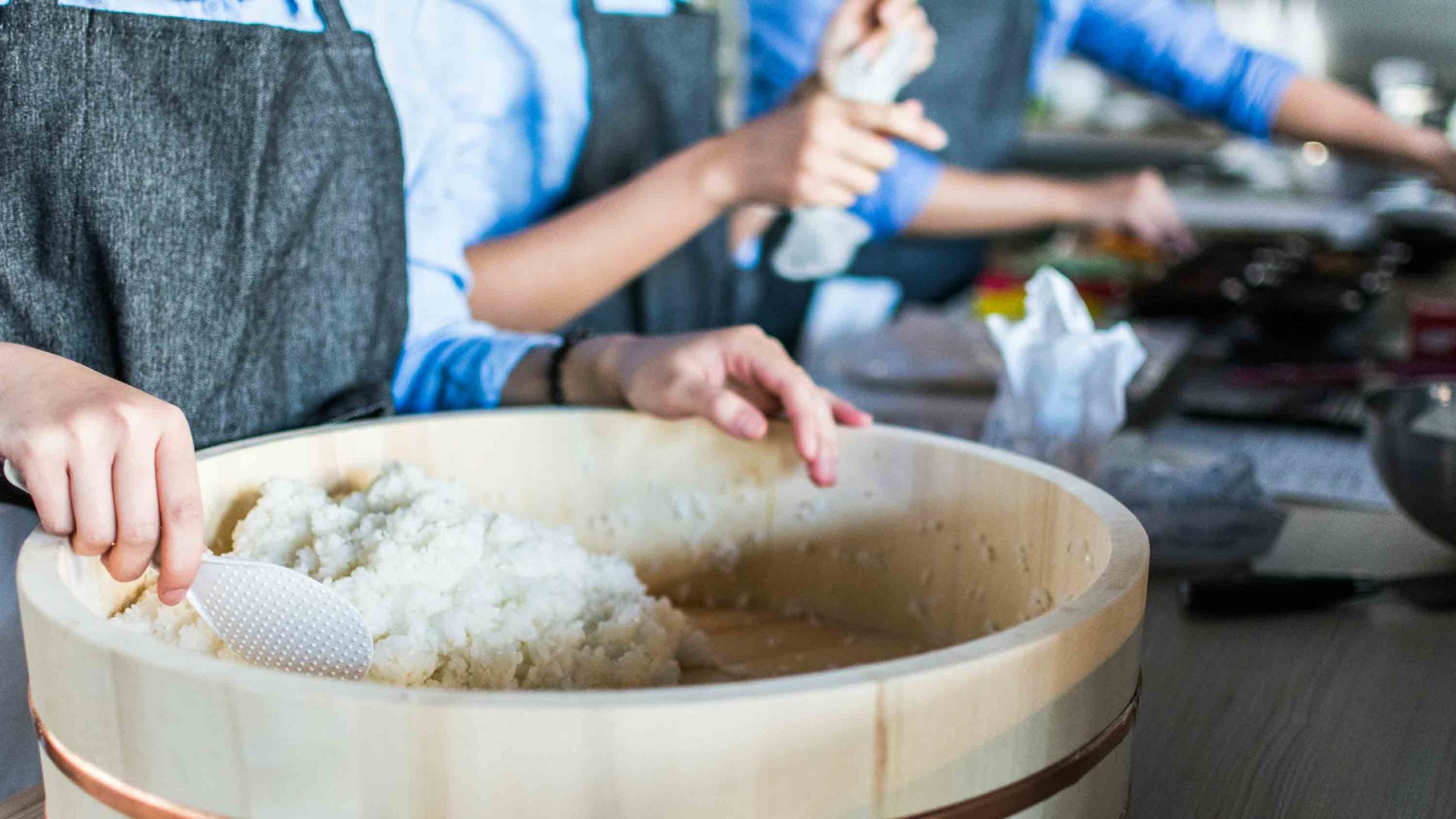 Three peoples arms are visible as they prepare food at a bench. One woman has her hands in a mixture in a big wooden bowl.