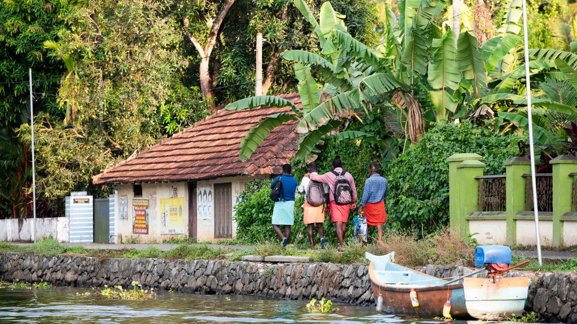 A group of friends with their arms around each other walk along the shore near a house and some trees.