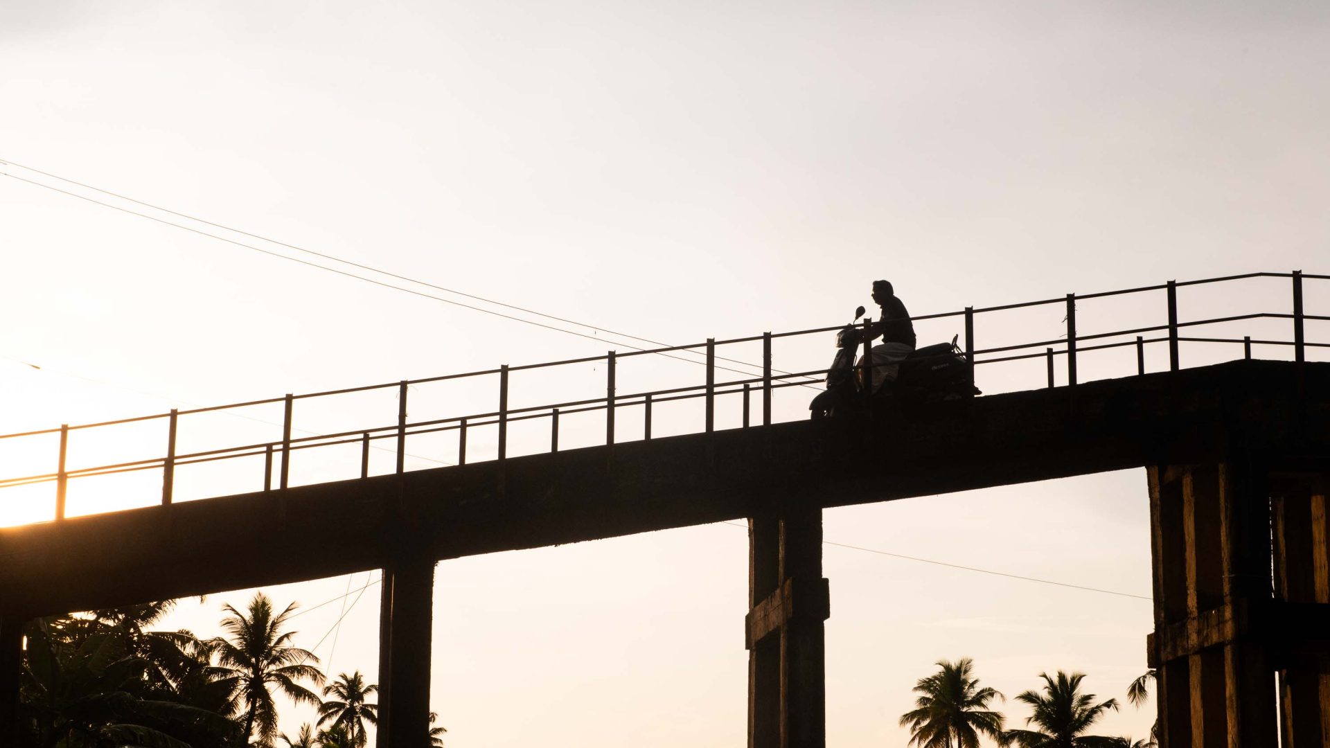 A silhouette of a person on a motorcycle riding down a small bridge.