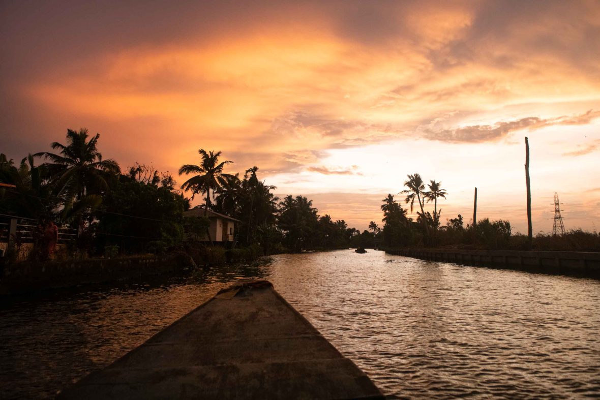 The bow of a small wooden boat moves through the water which has palm trees silhouetted on the shore and a vibrant orange sky.
