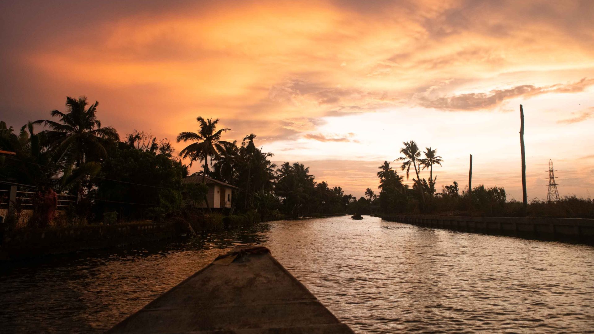 The bow of a small wooden boat moves through the water which has palm trees silhouetted on the shore and a vibrant orange sky.