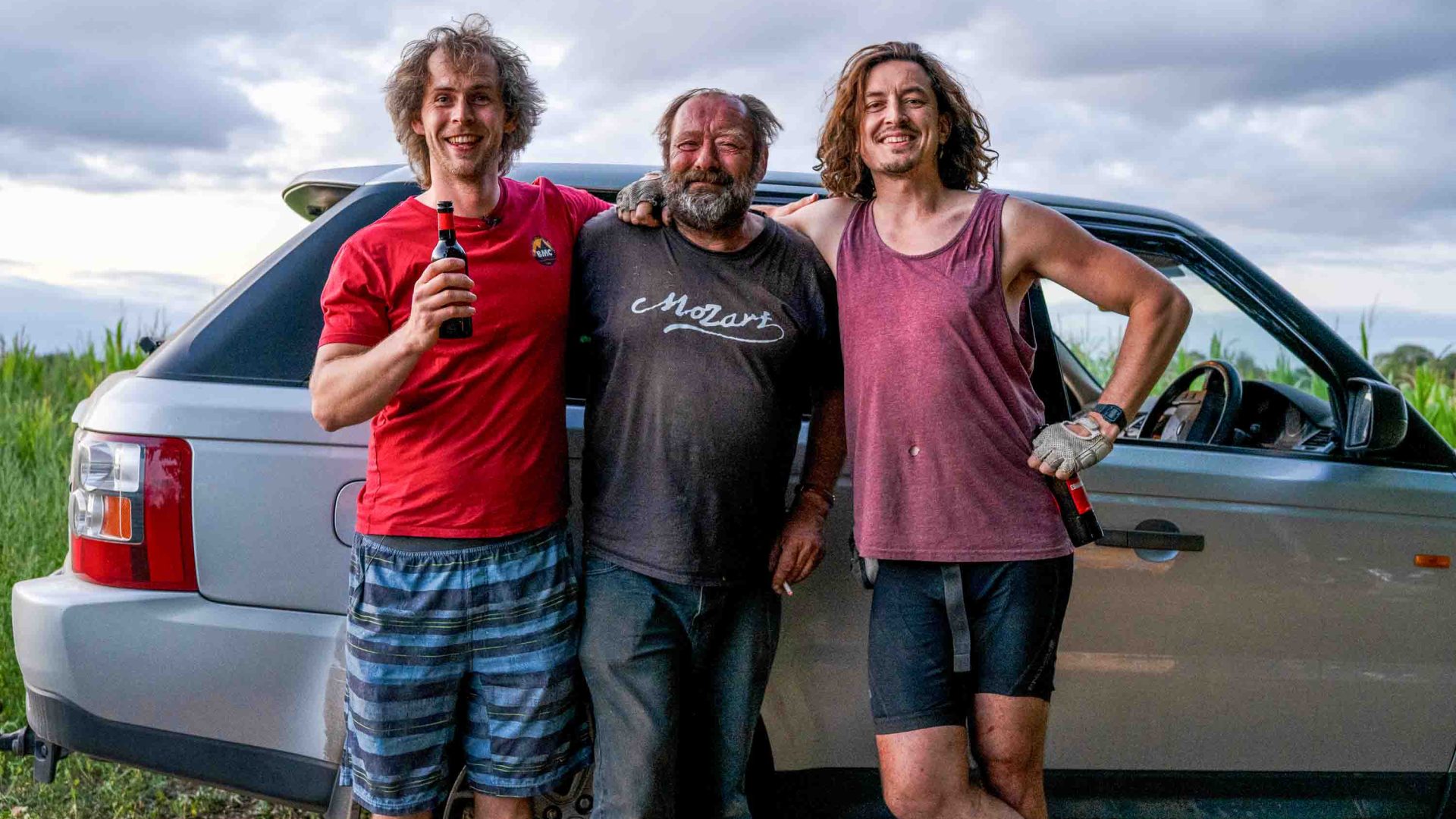Three men lean on a car and smile.