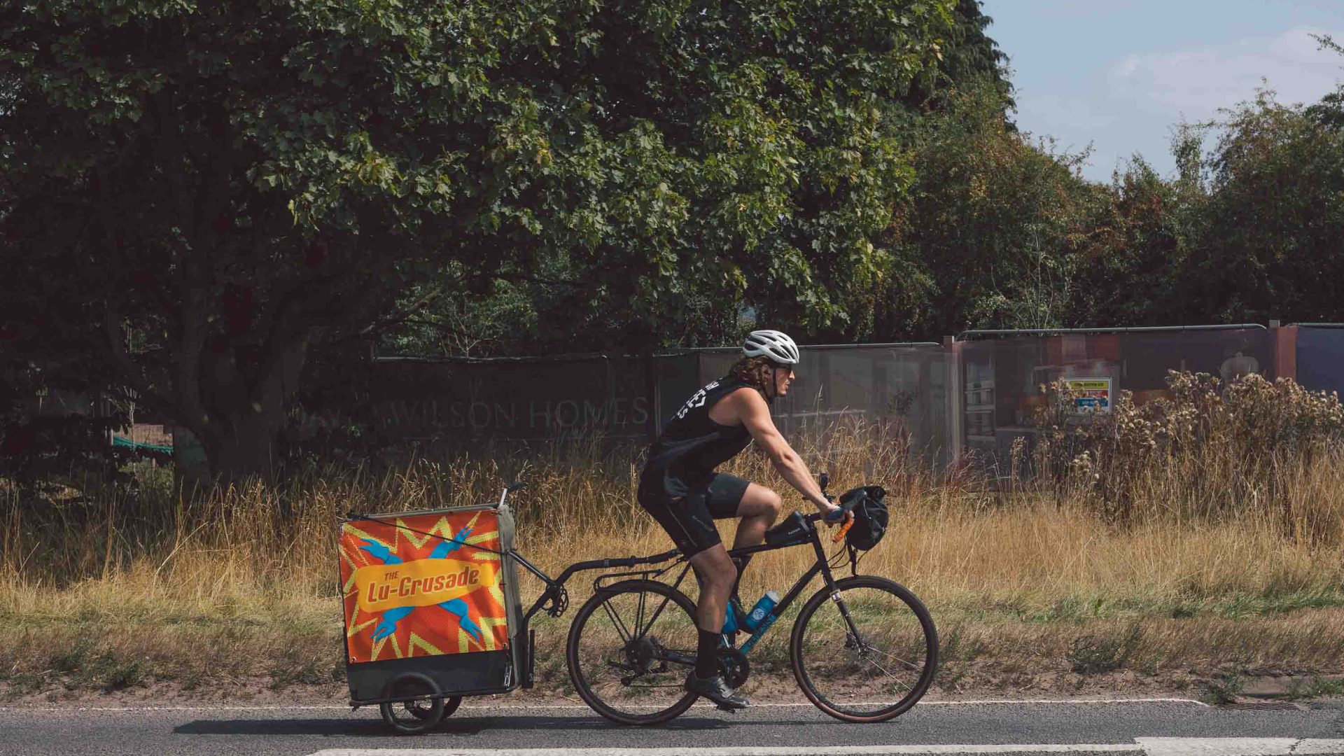 A man rides a bike, pulling a small square trailer behind him.