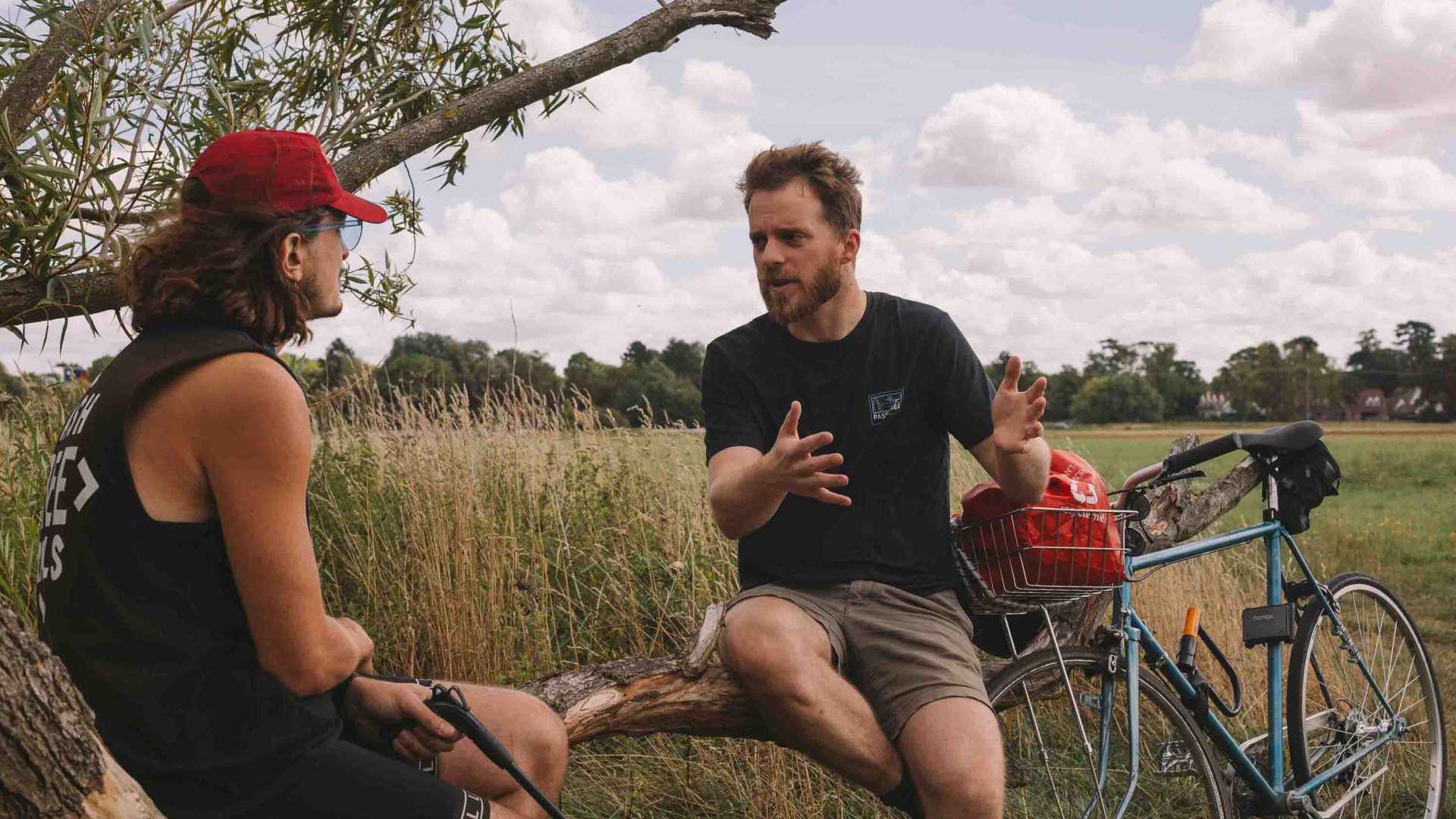 Two men sit on a branch in a field and talk.
