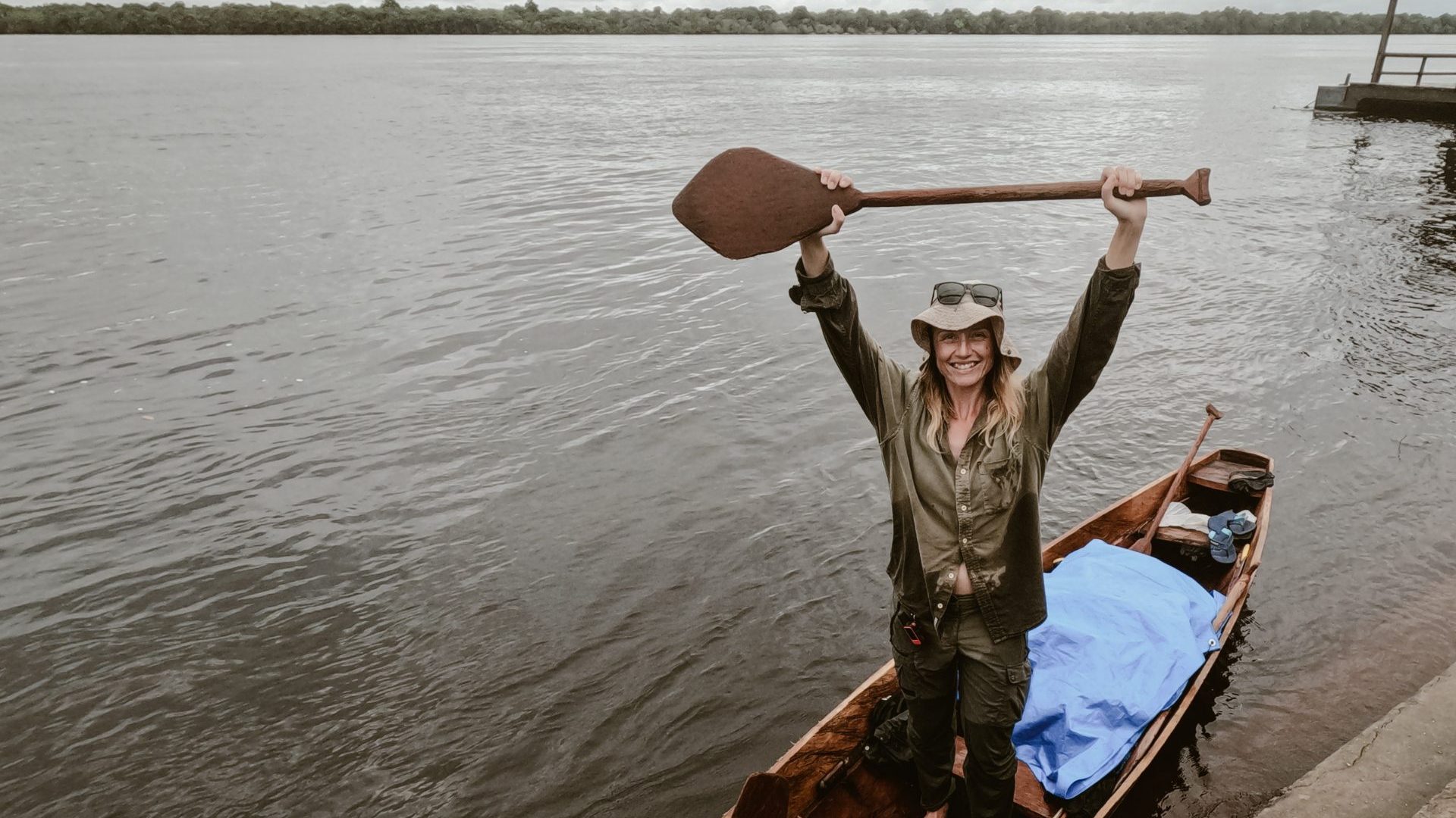 A woman standing in a canoe holds paddle up in air in triumph.