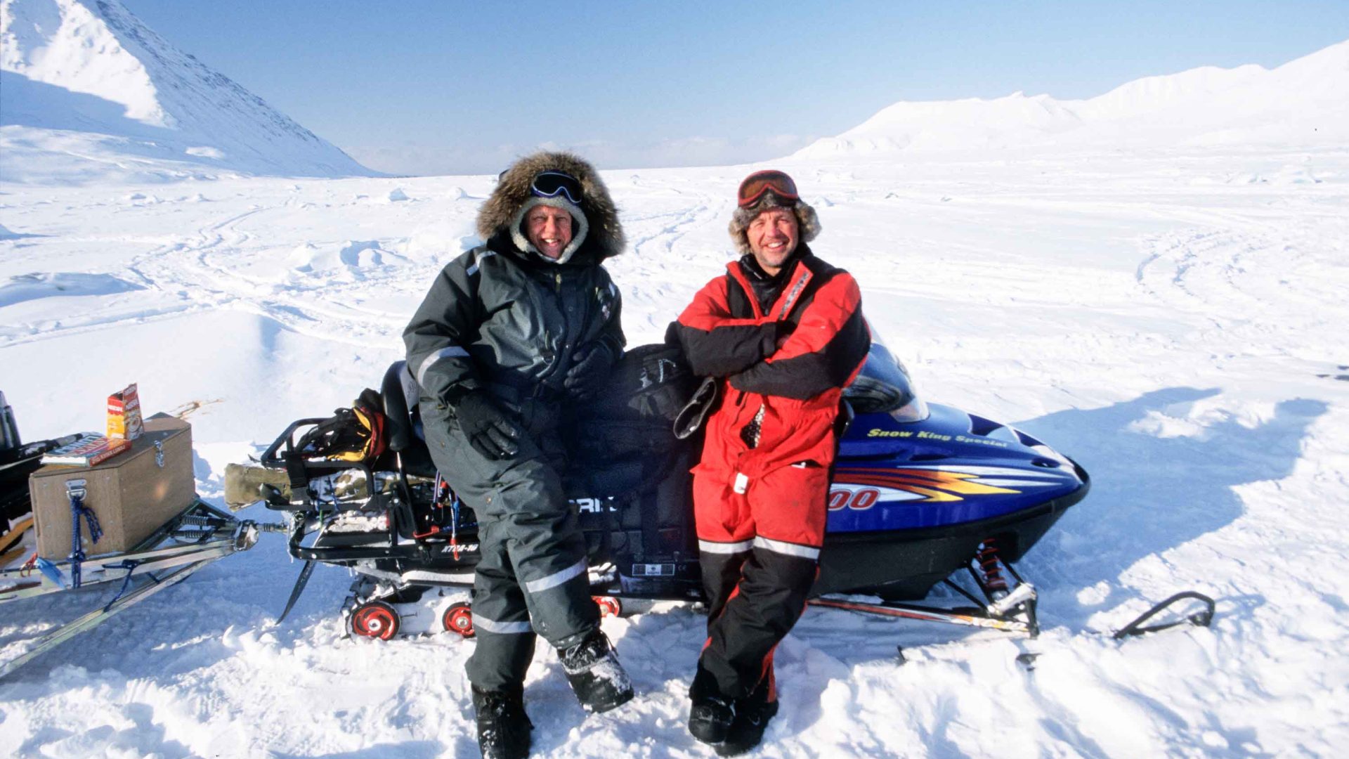 Two men lean against a ski mobile in the snow.