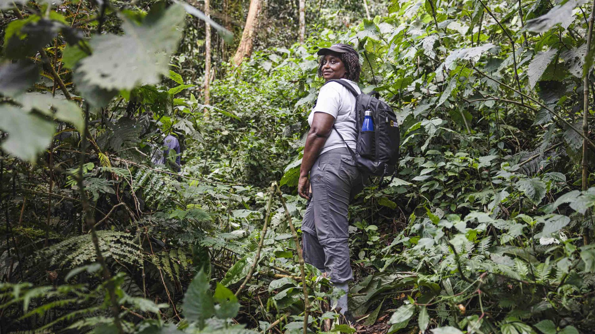 A woman in a hat stands in a jungle setting and smiles to the camera.