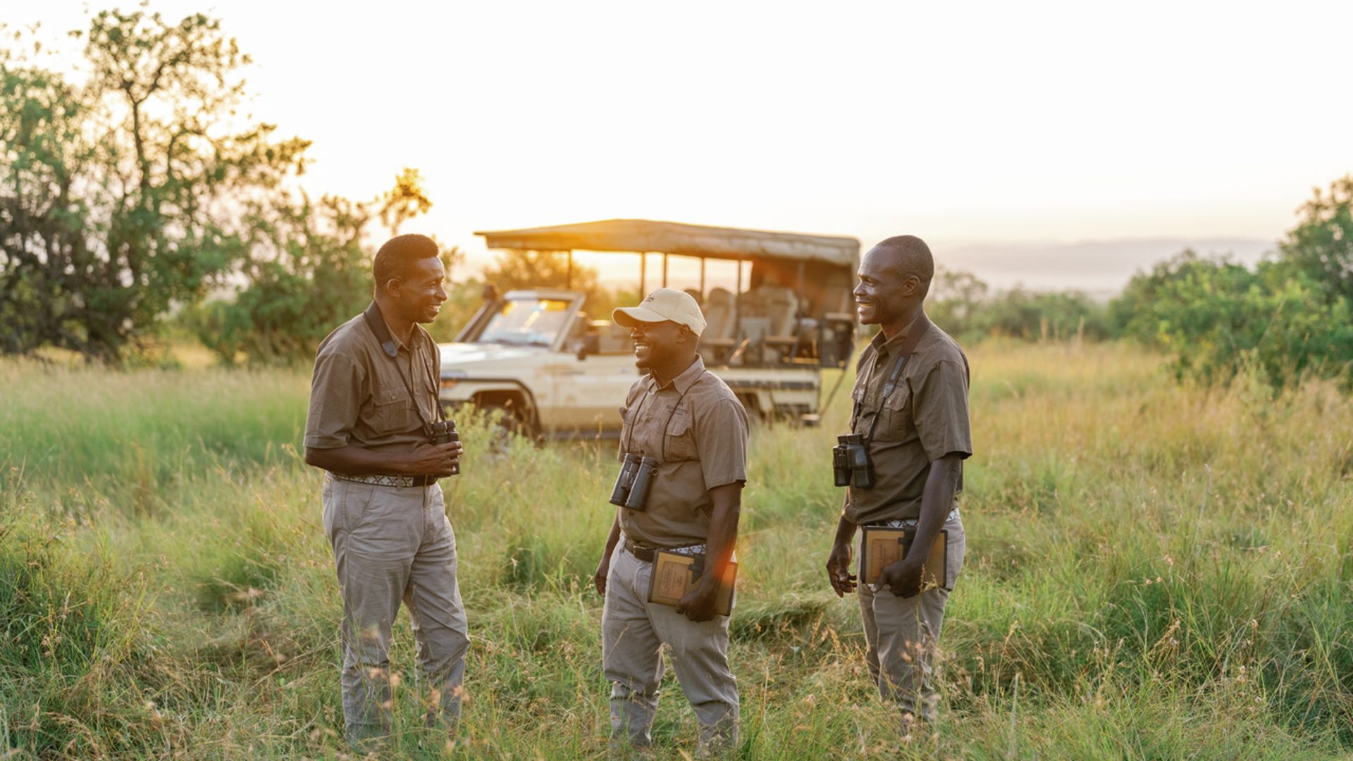 Three men in safari guide uniforms chat in grass with the safari vehicle in the distance behind them.