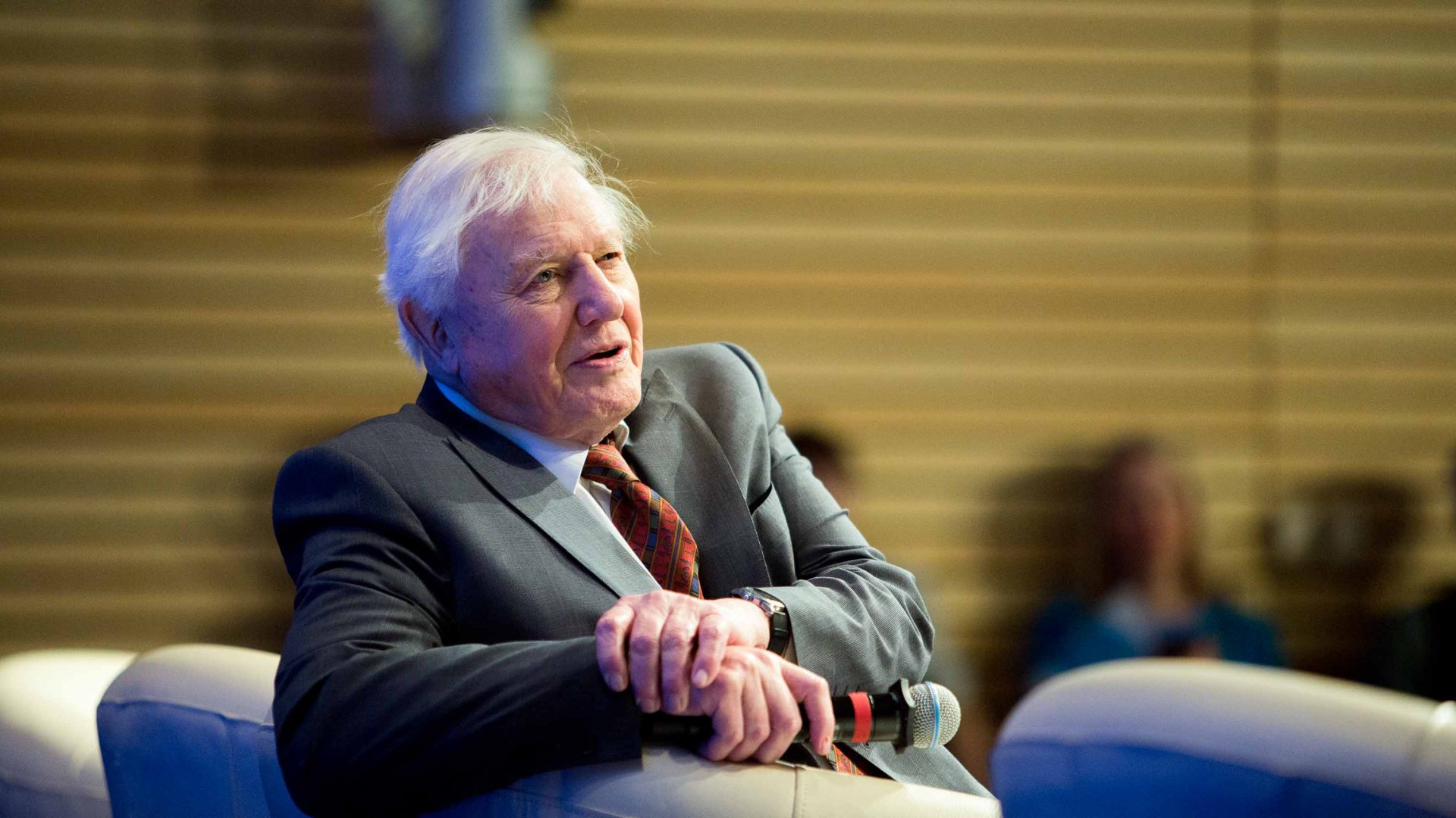 A man holding a microphone looks away. He is seated.