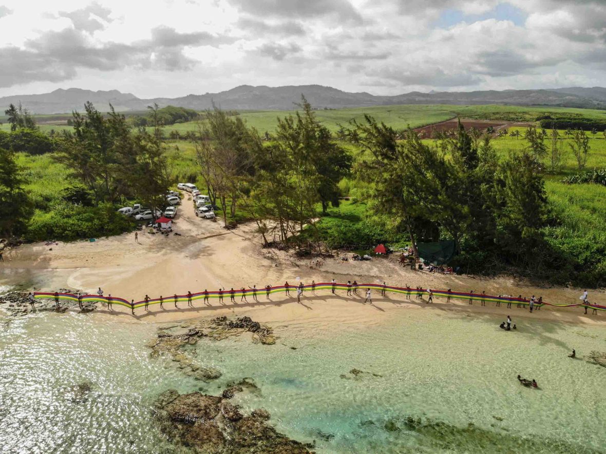 A drone shot of people holding a long ribbon on a beach surrounded by forest.