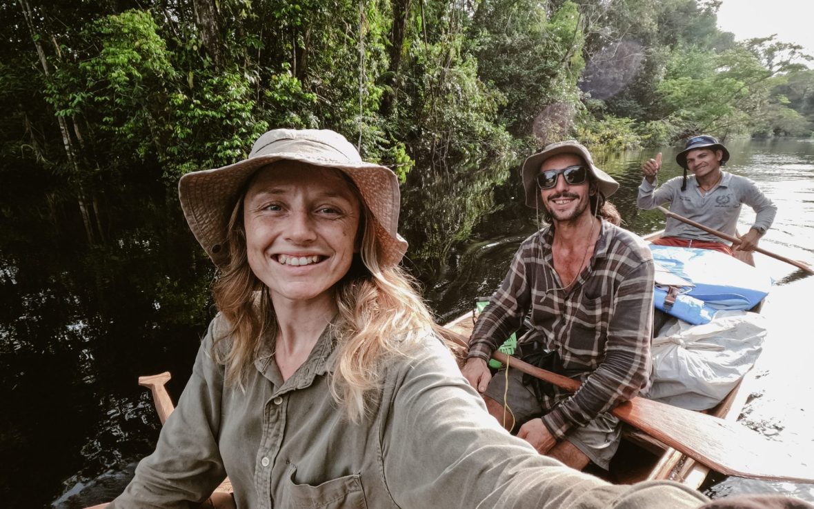 A blonde woman and two men smile from a dugout canoe floating on dark river water