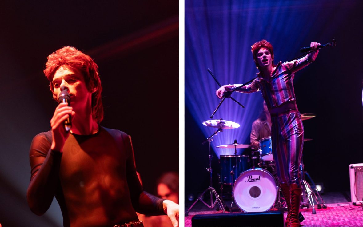 Man wearing red wig sings into microphone and dances in front of drum set.