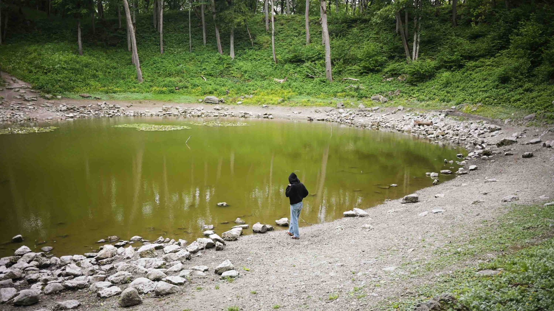 A person in black stands alongside a green pool of water, which is a crater.