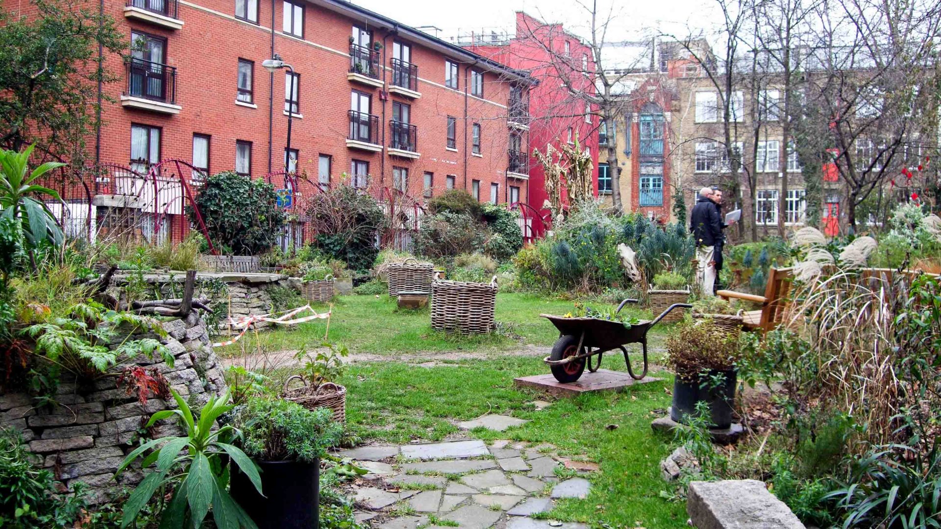 A red brick apartment building stands alongside a community garden with paths, wheelbarrows and places to sit.