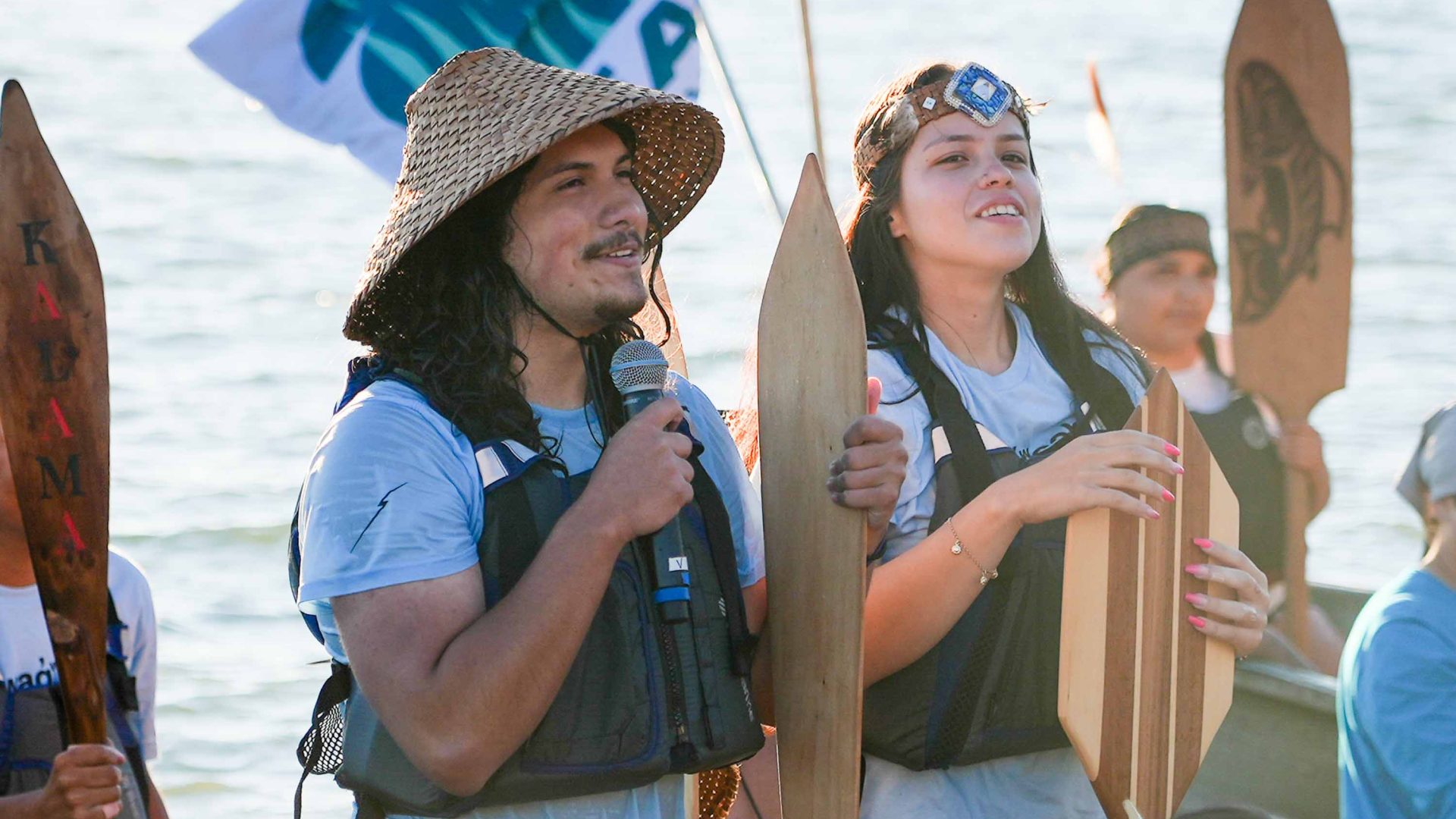 Two people in traditional First Nations head wear hold wooden oars and speak int a microphone.