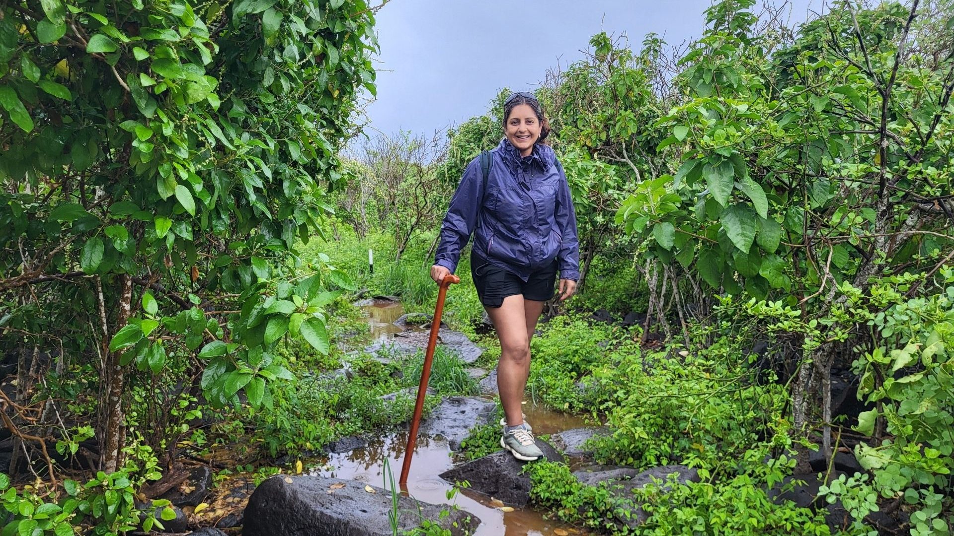 A woman in shorts and jacket hiking in the rain against a lush green backdrop