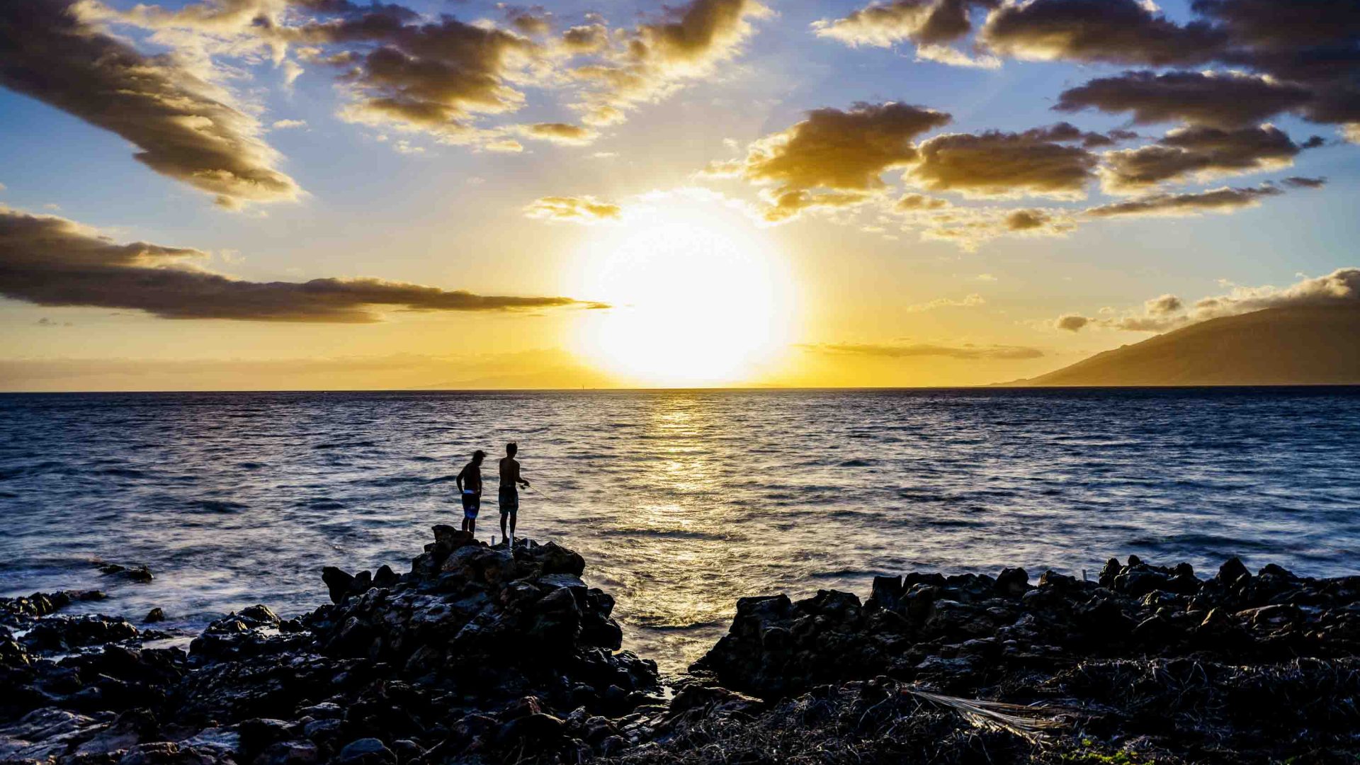 Two people are silhouetted on a rock, fishing at sunset.