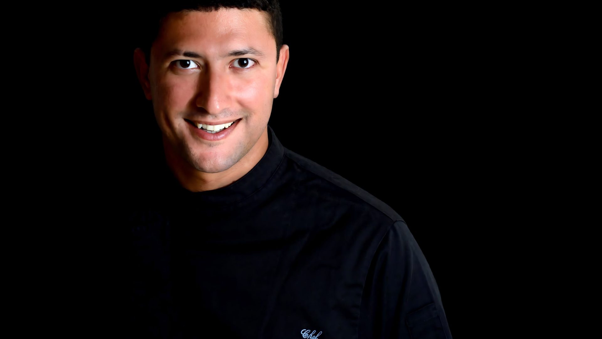 A man in black smiles to camera. He is against a black background.