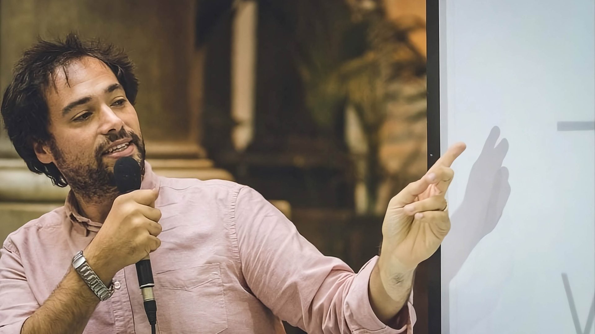A man with a microphone points to a whiteboard.