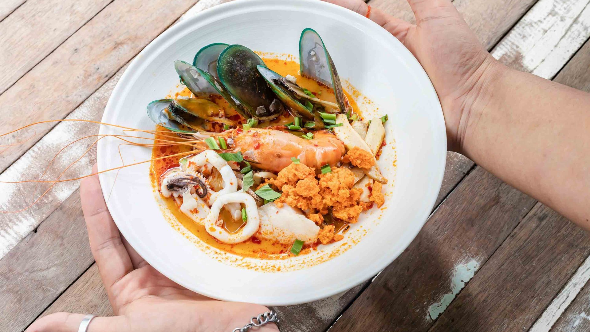 Hands hold a plate of tom yum soup with lots of fresh seafood in it.