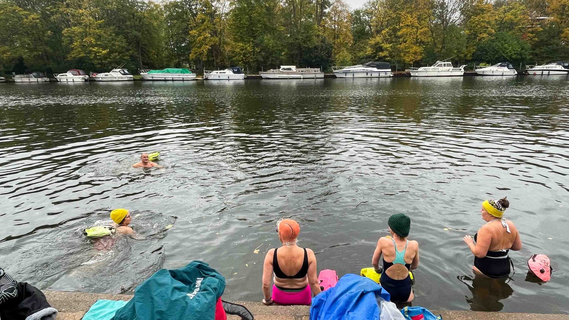 A group of five people swim in a river. their belongings are pictured on the shore in the foreground.