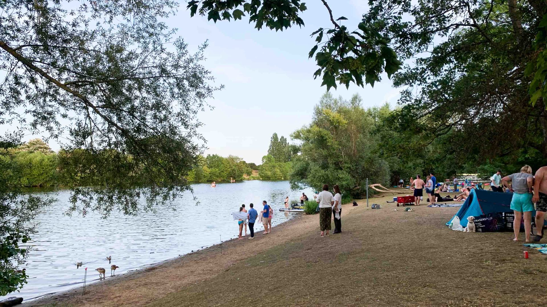 A number of people stand and sit on the sandy banks of the river, framed by trees.
