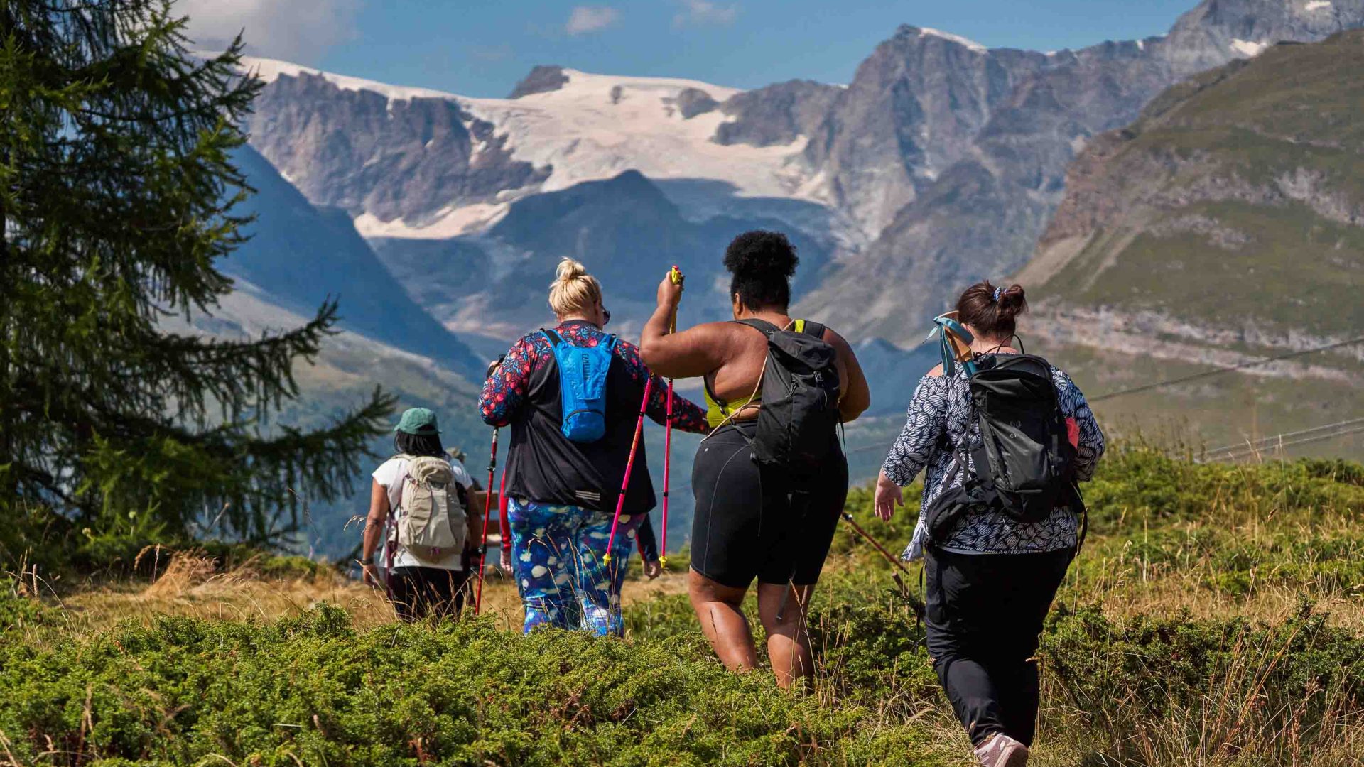 A group of women hike through a valley with mountains in the background. Their backs are to the camera.