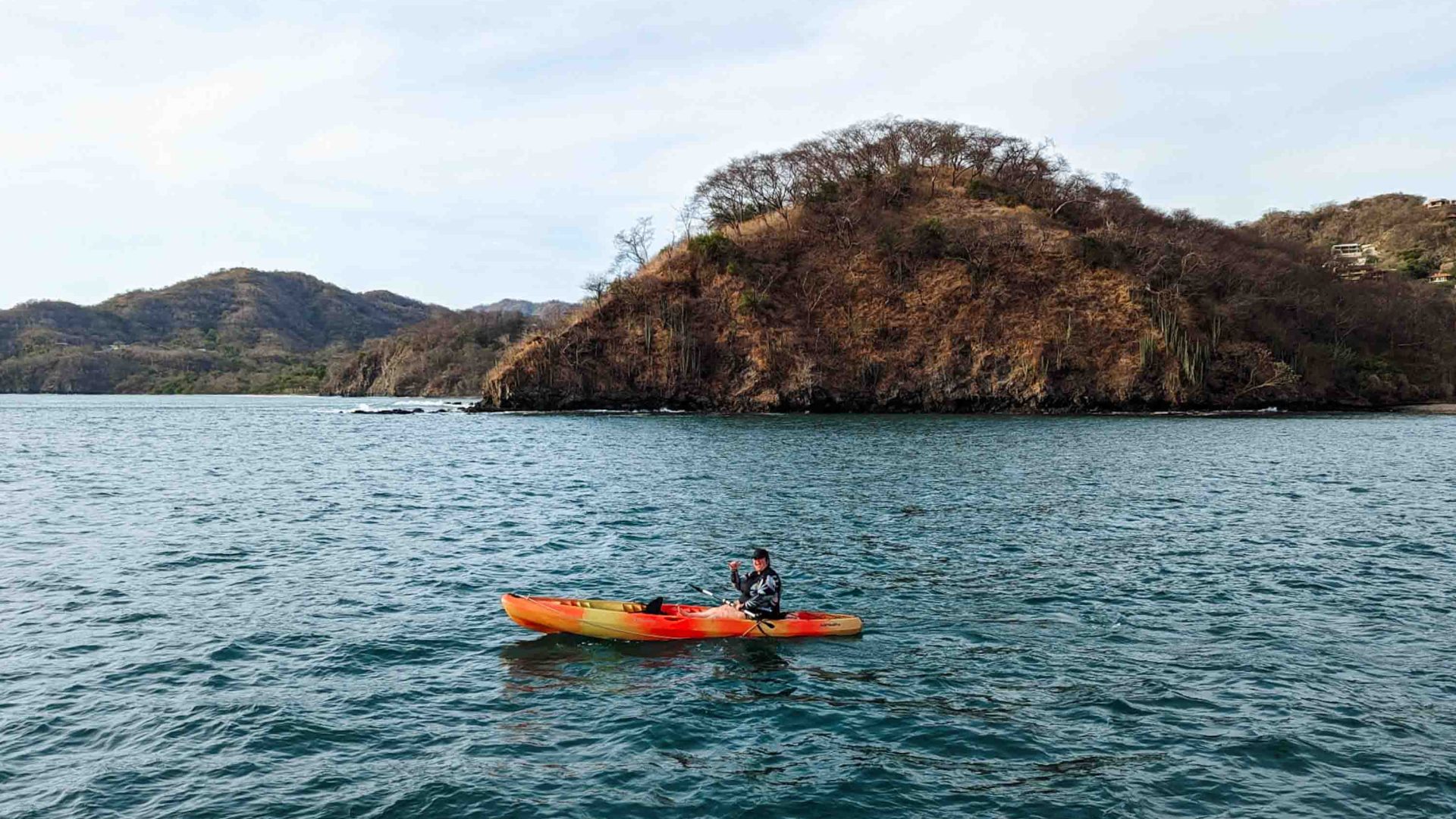 A woman in a red kayak in flat water with a tree covered hill behind her.