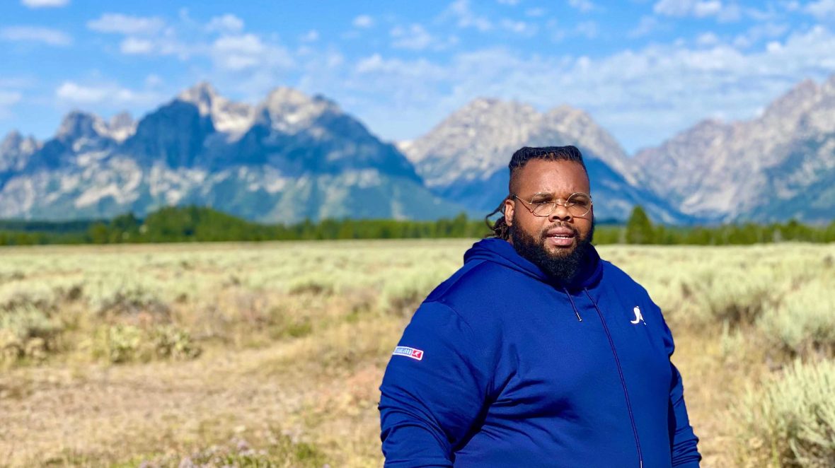 A man in blue looks serious as he stands in front of a vista of mountains and fields.