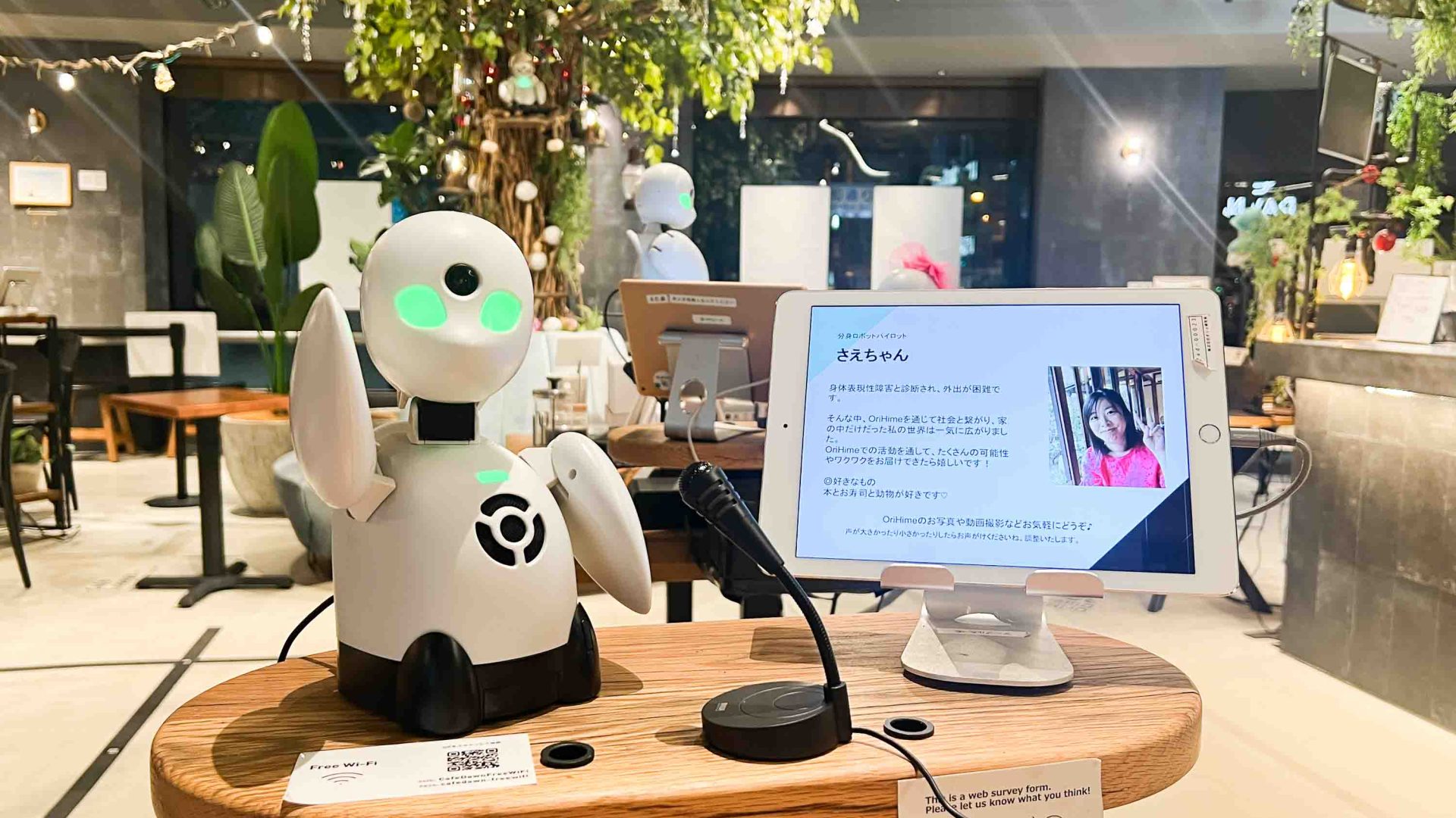 A robot with one of the pilots that controls it remotely on a screen to its side. The robot has its hand in the air.