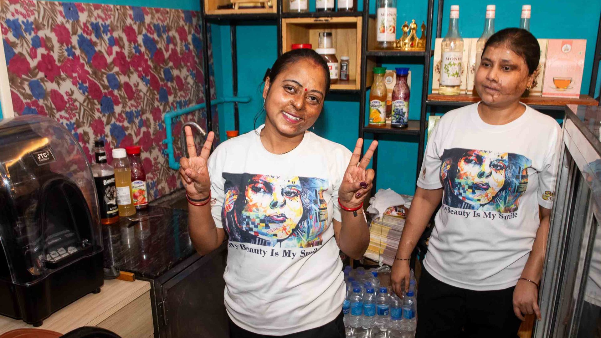 Two women smile. They wear matching t-shorts and both have scarring from acid attacks.