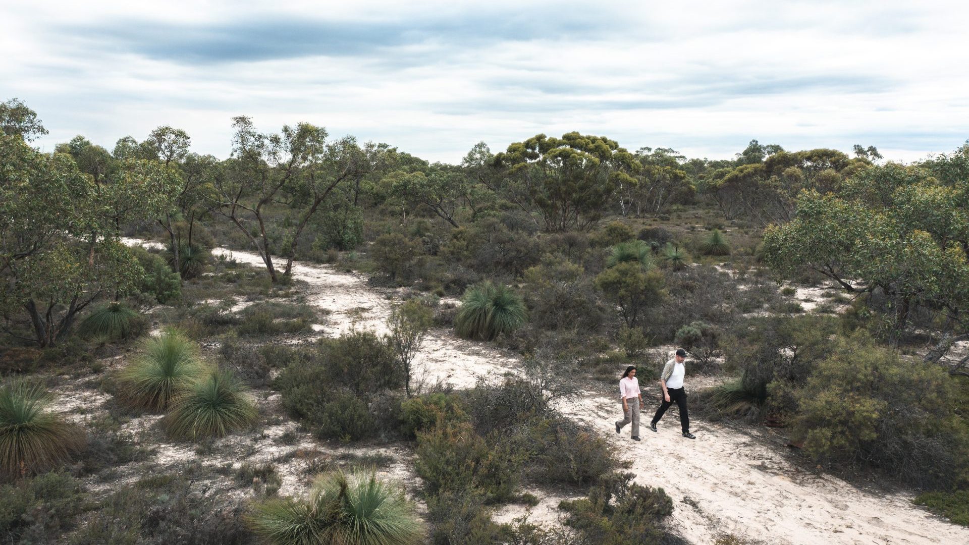 Man and woman walk down sandy path between green bushes.