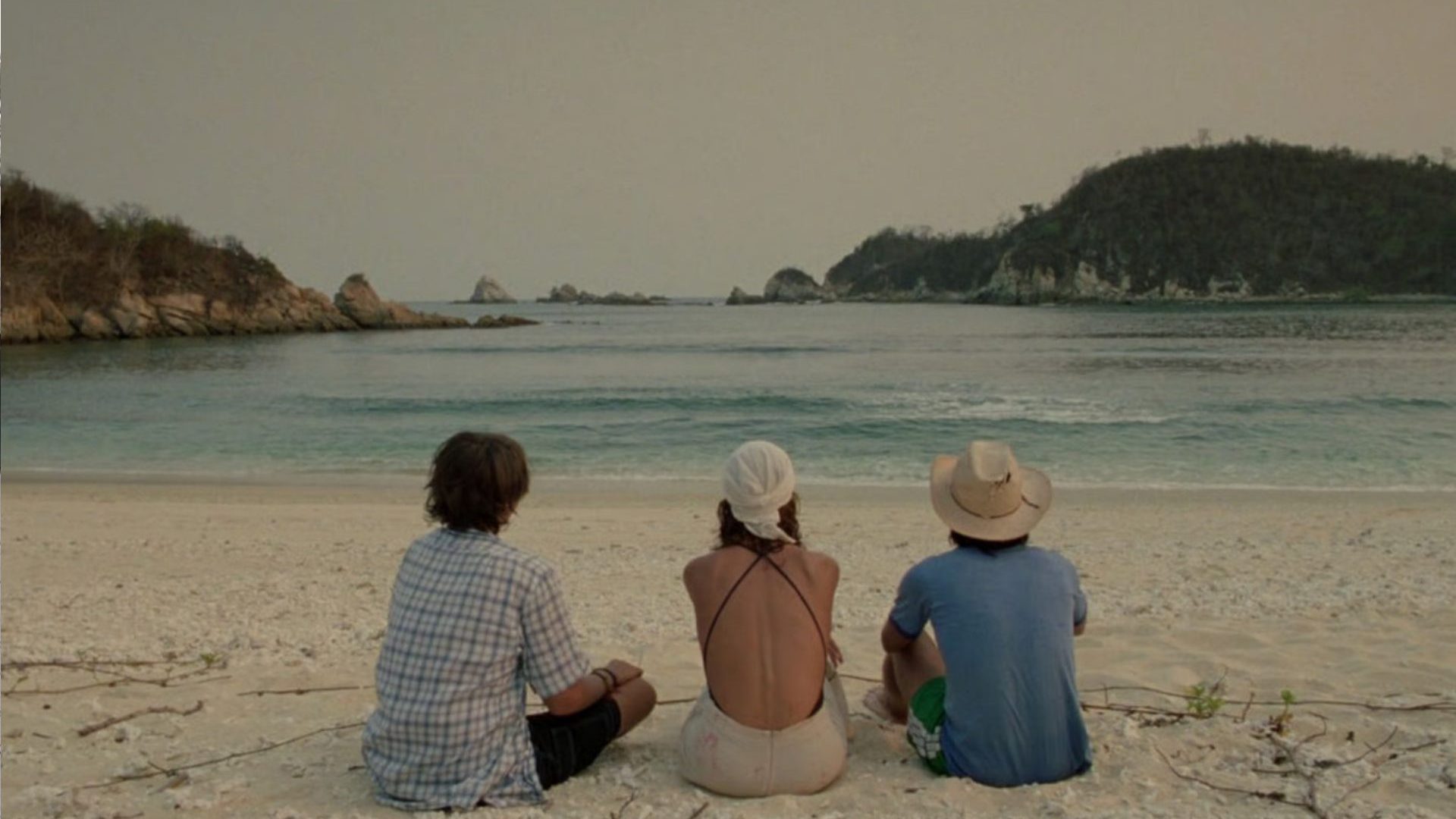 Three people sit on beach facing the water.