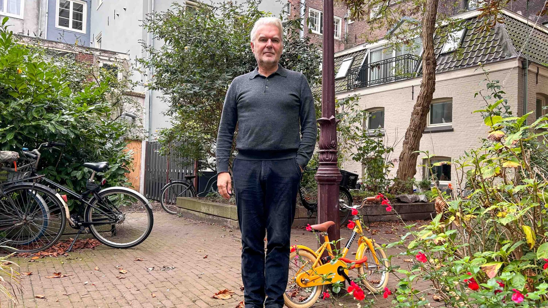 A man stands outside his home. There is a bike on the ground near him.