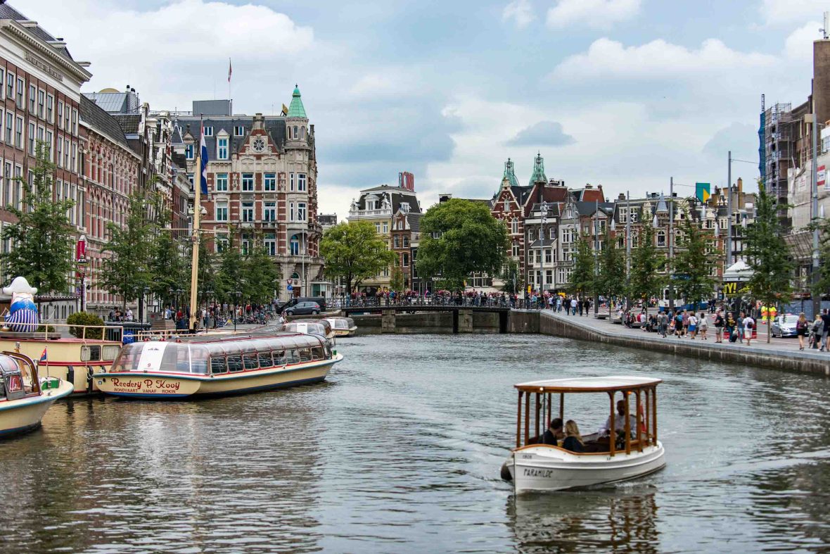 Boats travel up a canal with a bridge over it and houses on the sides.