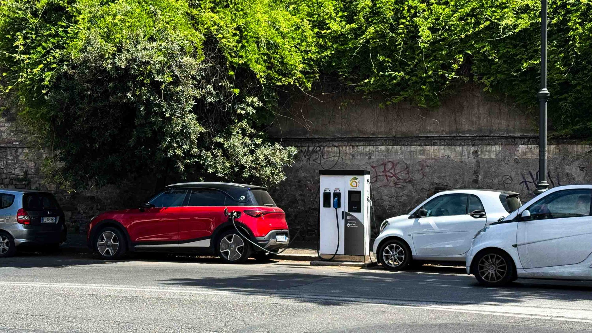 A red electric vehicle charging at a white charging point in the street.