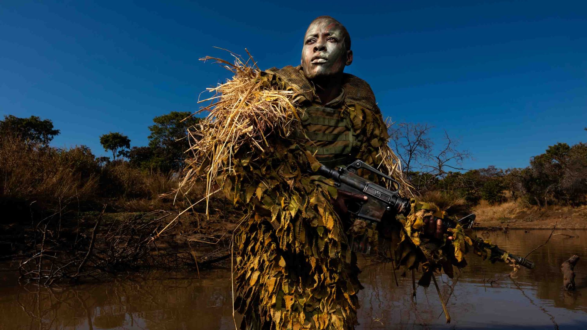 A woman is camouflaged behind sticks and carries a gun.