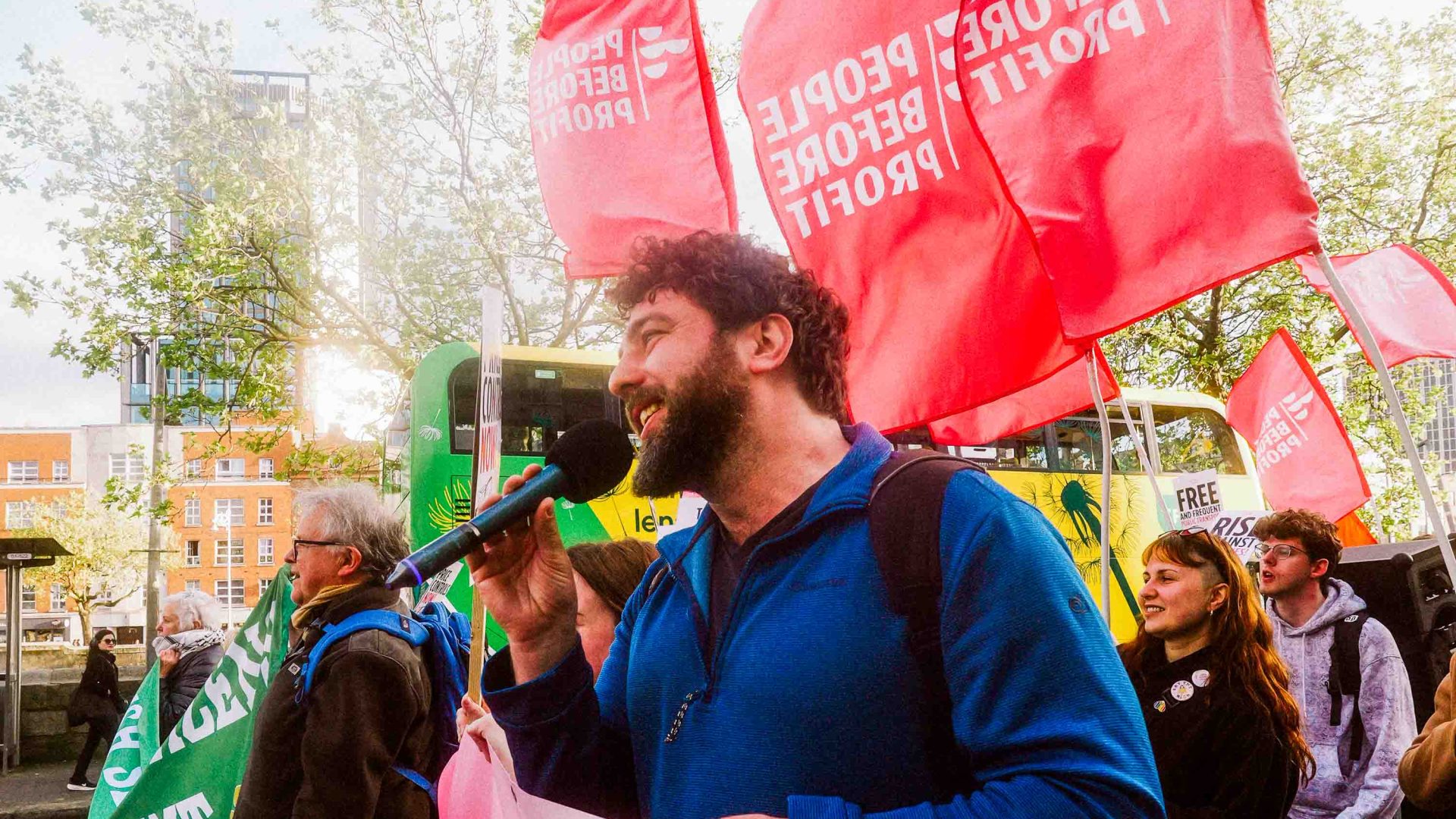 A man speaks into a microphone. There are red flags and crowds of people behind him.