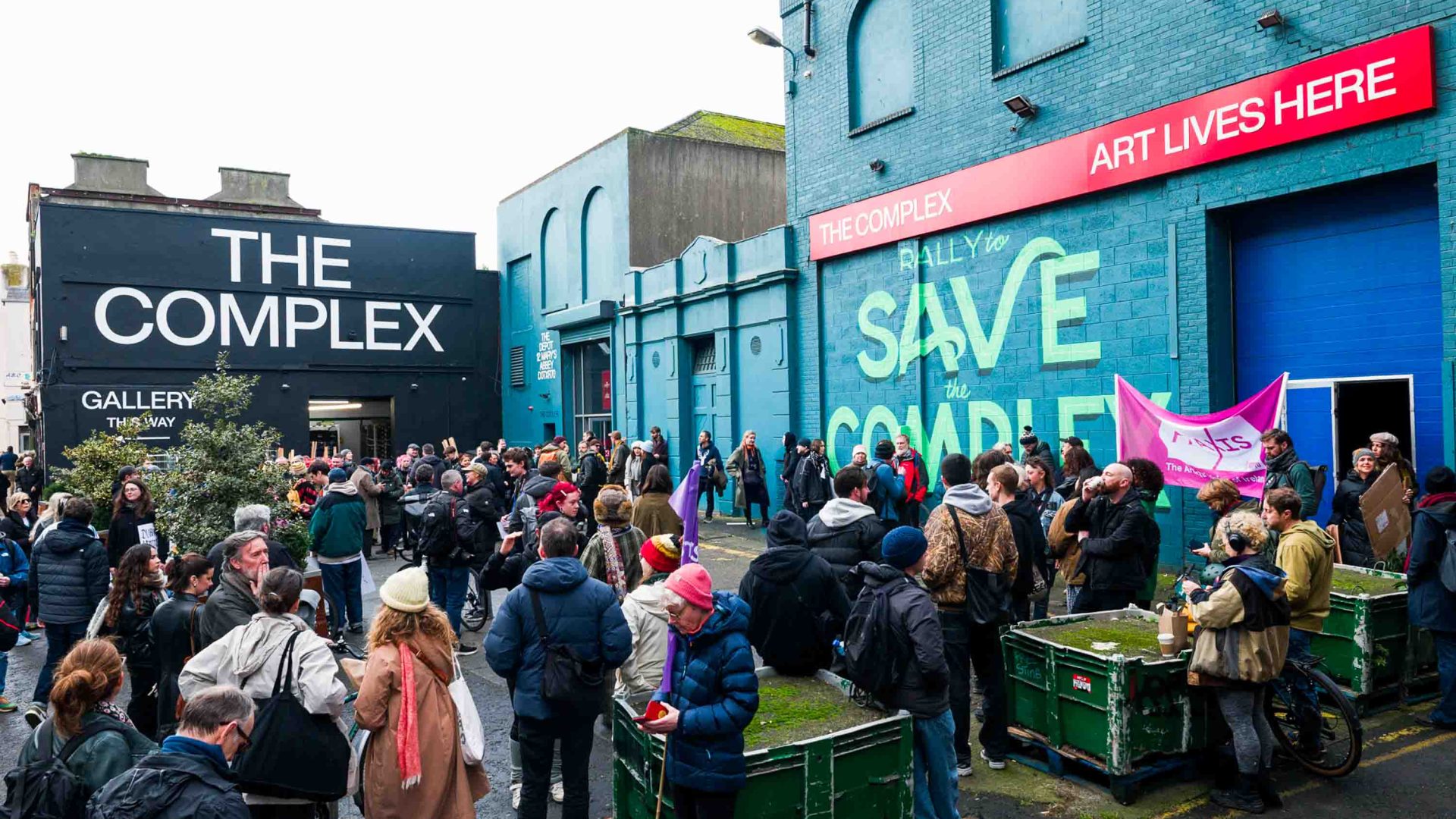Lots of people mill outside of a black and green building with the words "The Complex" painted on its walls.