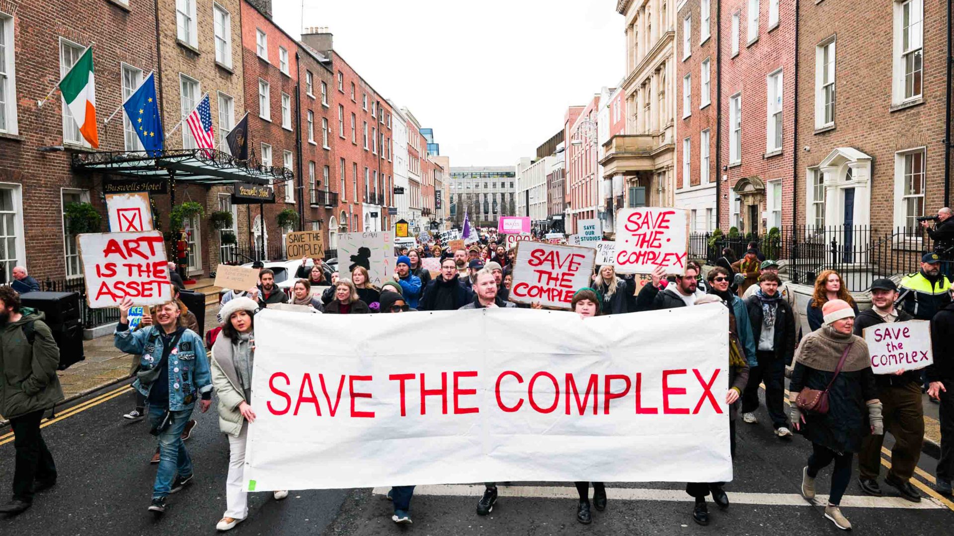A number of protestors in the street. they hold a big banner that reads "Save the Complex".