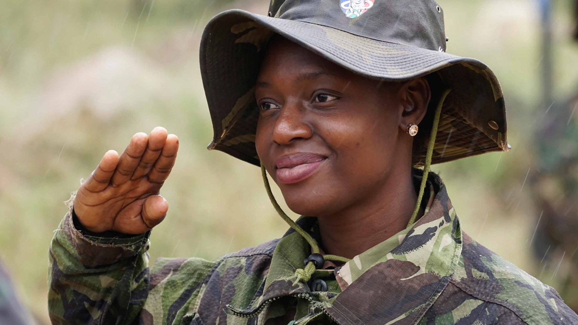 A woman in army fatigues and a hat does a salute while rain falls around her.