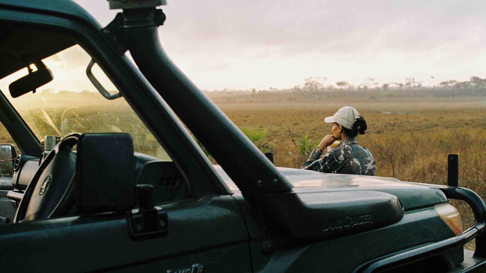 A woman stands alongside a jeep looking out at some African plains.