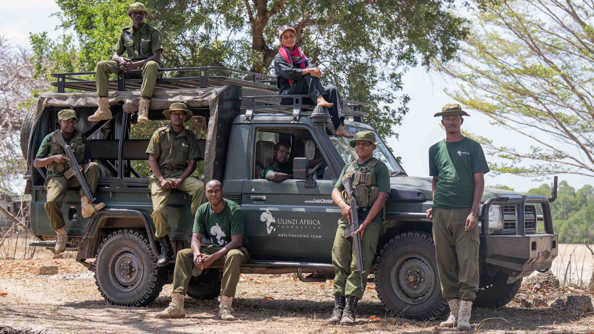 A woman sits on the top of her jeep. She is surrounded by a number of male rangers, and they all pose for a photo.