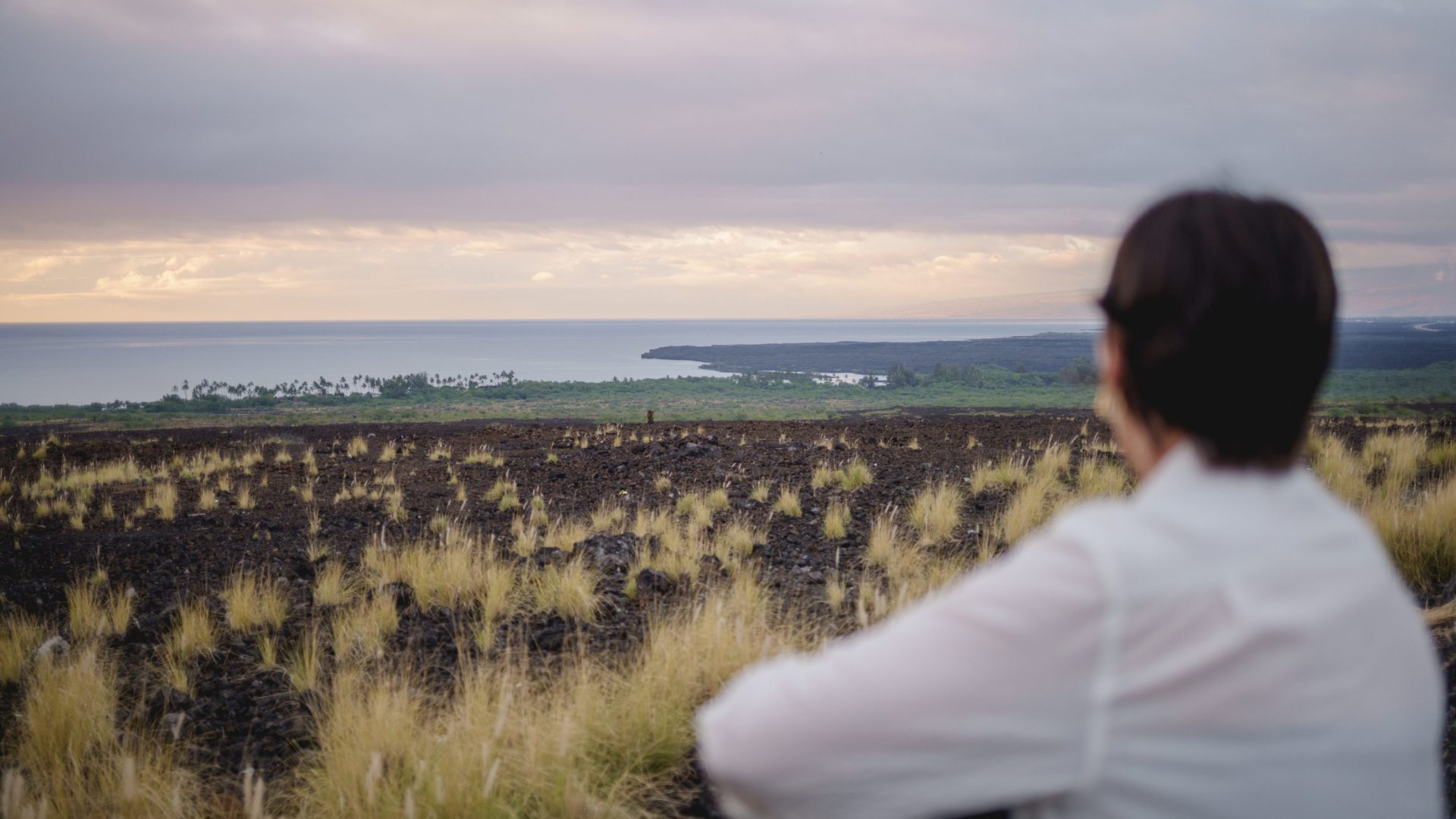 Woman looks out across black lava landscape that leads to the sea.