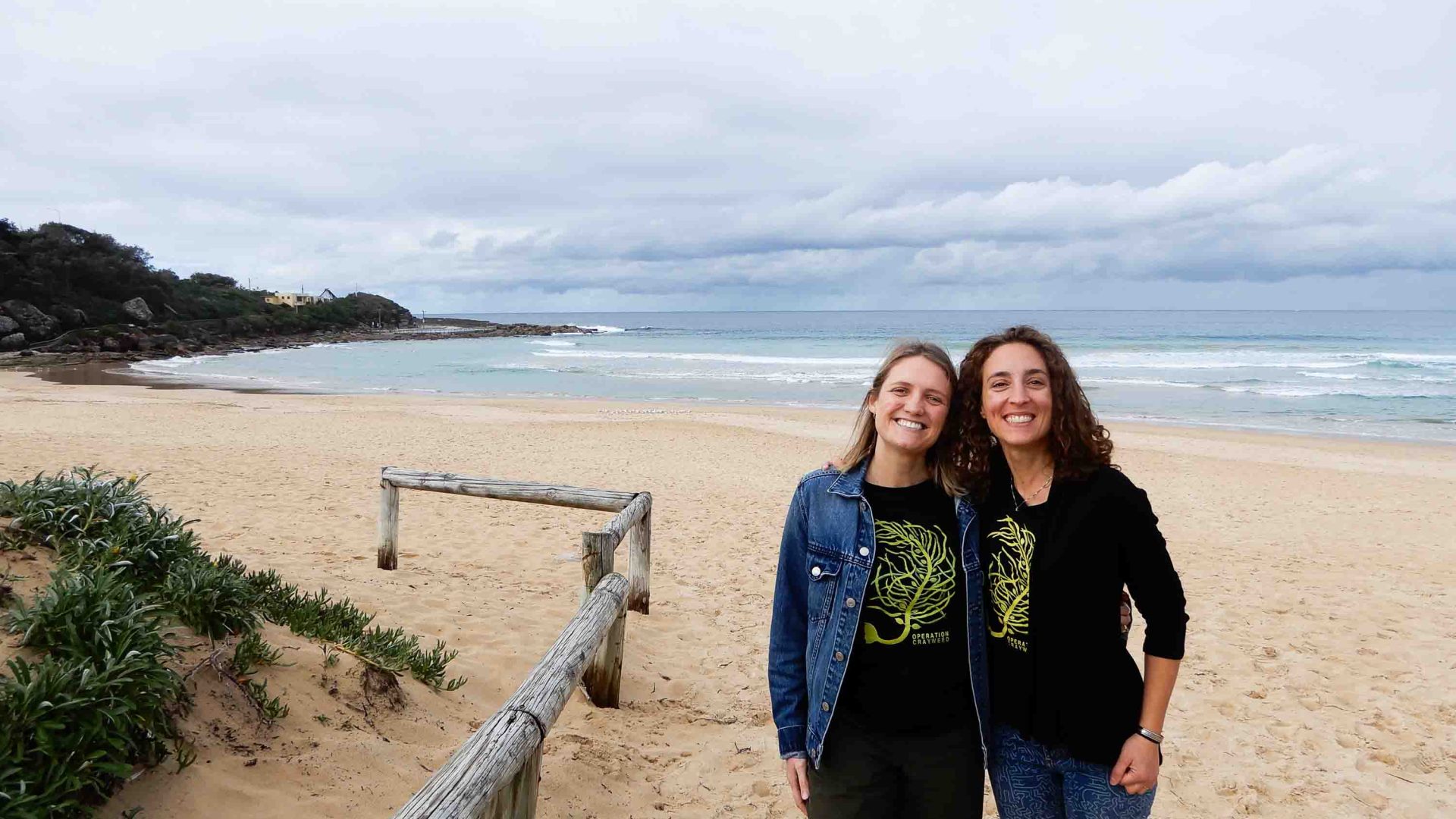 Two women smile while standing in front of a beach.